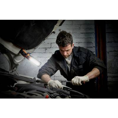 A mechanic is repairing a car's engine with tools in a garage. A lamp is illuminating the area.