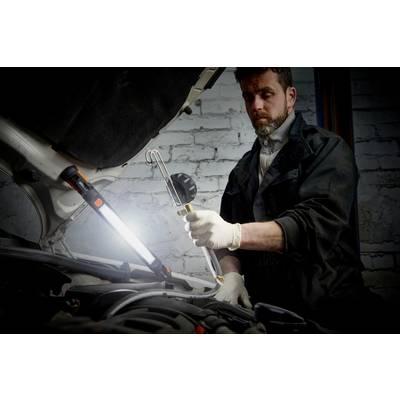 A mechanic inspects the engine of a car in a workshop. He is wearing gloves and holding a work light, which is directed at the engine.