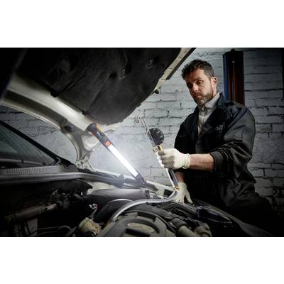 Mechanic repairing an engine, standing beside an open car with a work lamp. Workshop environment, with a brick wall in the background.