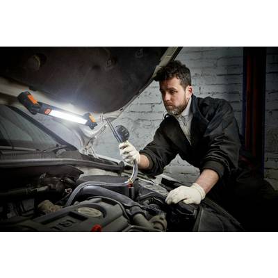 A mechanic in overalls is checking the engine of a car in a workshop. He is holding a diagnostic device while a work light illuminates the scene.