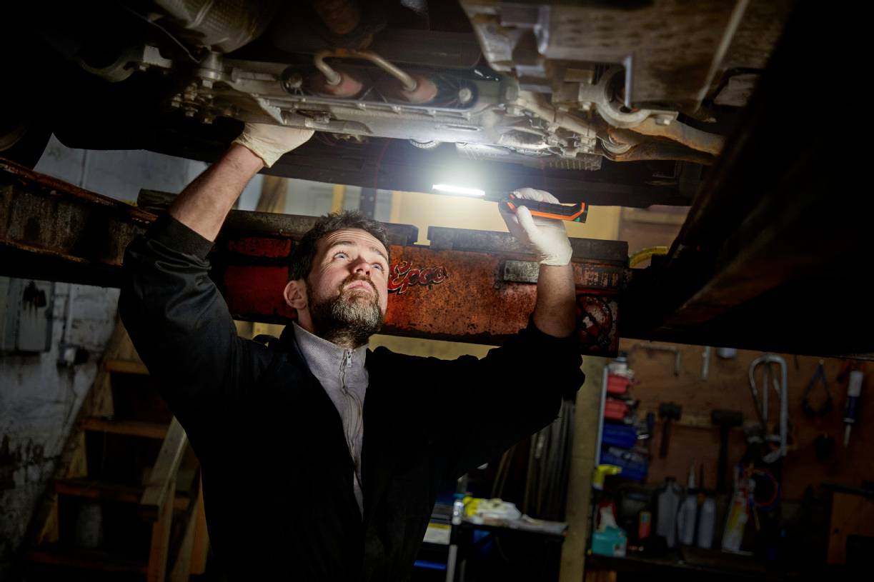 A mechanic is working underneath a car in a garage. He is using a lamp to illuminate the underside of the vehicle.
