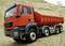 A red lorry stands on gravelly ground in front of a sand mound. A logo is visible in the background.
