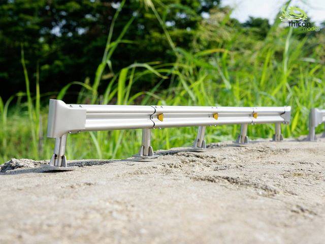 A small metal model fence stands on a stony surface, with green grass and vegetation visible in the background.