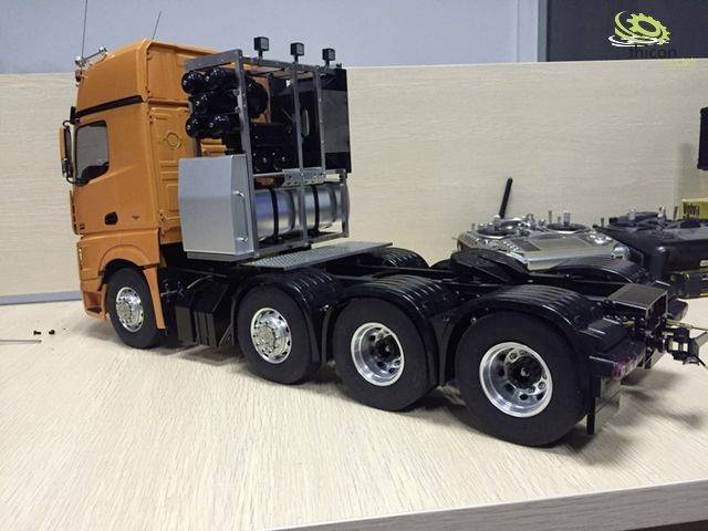 A model lorry in orange with a black trailer is sitting on a table. The lorry has seven wheels and some technical details are visible.