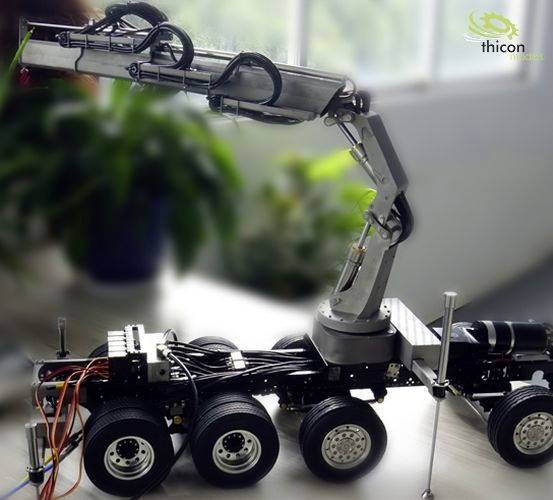 A model vehicle with six wheels and a mechanical arm sits on a table. Plants can be seen in the background.