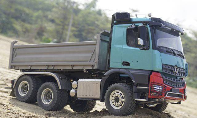 A green lorry with a load bed drives over sandy terrain, surrounded by trees.