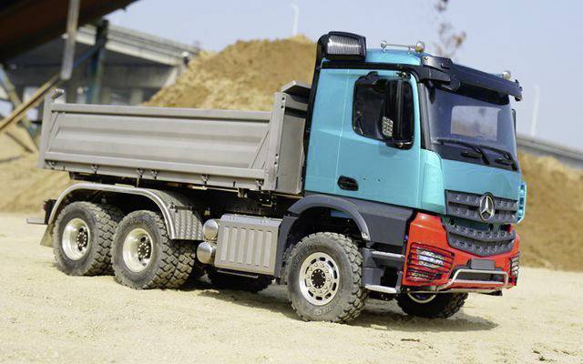 Lorry on a construction site, parked in sand in front of an earth mound. The vehicle is turquoise with a red bumper and has six wheels.