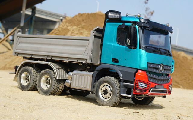 Lorry on a construction site, parked in sand in front of an earth mound. The vehicle is turquoise with a red bumper and has six wheels.