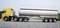 A yellow lorry with a silver tanker trailer is parked on a paved road. Trees are visible in the background.