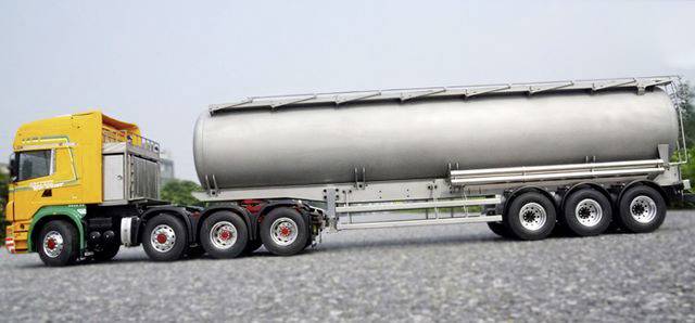 A silver tanker truck with a yellow cab is parked on a grey gravel area, surrounded by a green background.