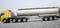 A silver tanker truck with a yellow cab is parked on a grey gravel area, surrounded by a green background.
