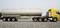 A yellow tanker lorry on a grey road. It has six wheels on the tractor unit and three on the trailer. Clear, bright surroundings.