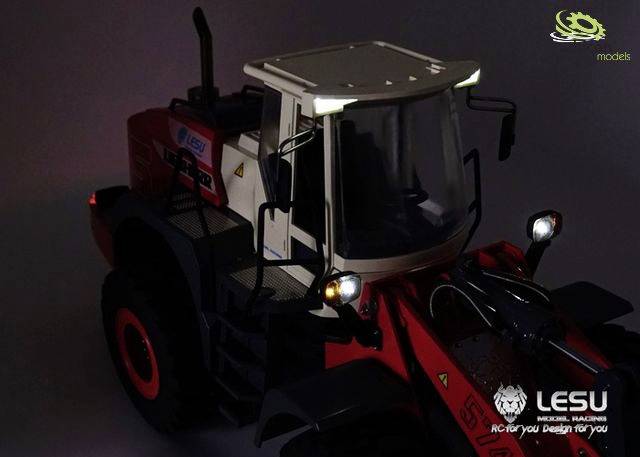 A wheeled loader model with red and black design stands in the foreground. It has a cabin with illuminated headlights.