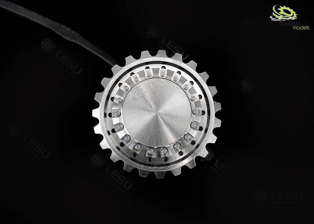 A metallic gear wheel against a black background, detailed view. The precisely machined surface reveals multiple small perforations.