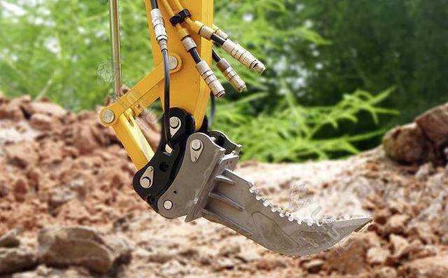Close-up of a yellow excavator arm with a serrated bucket tip, working in an earthy terrain, surrounded by green vegetation.