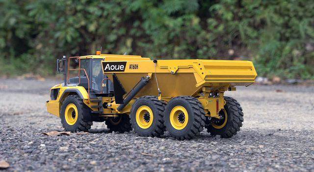 A yellow dump truck sits on a gravel surface, surrounded by green vegetation. Model vehicle with meticulous detail.
