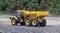 A yellow dump truck sits on a gravel surface, surrounded by green vegetation. Model vehicle with meticulous detail.