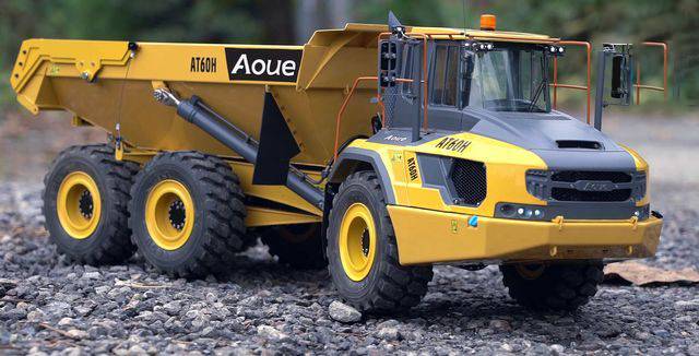 A yellow dump truck with the inscription 'AT60H' is driving on a gravel road. The background shows a blurred forest.