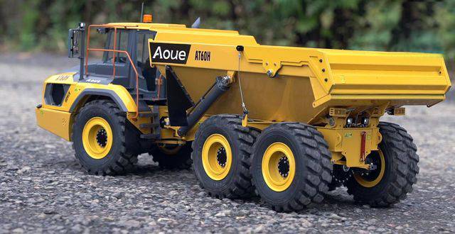 A yellow dump truck with the inscription 'Aoue AT61N' is parked on a gravel surface. The background is blurred with trees.