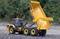 A yellow toy tipper truck on a gravel surface with its cargo area raised, surrounded by green vegetation in the background.
