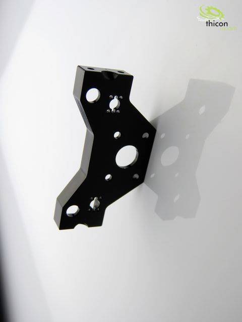 Black metal component in front of a white wall, showing recesses and holes, with a shadow. A green logo in the top right corner.