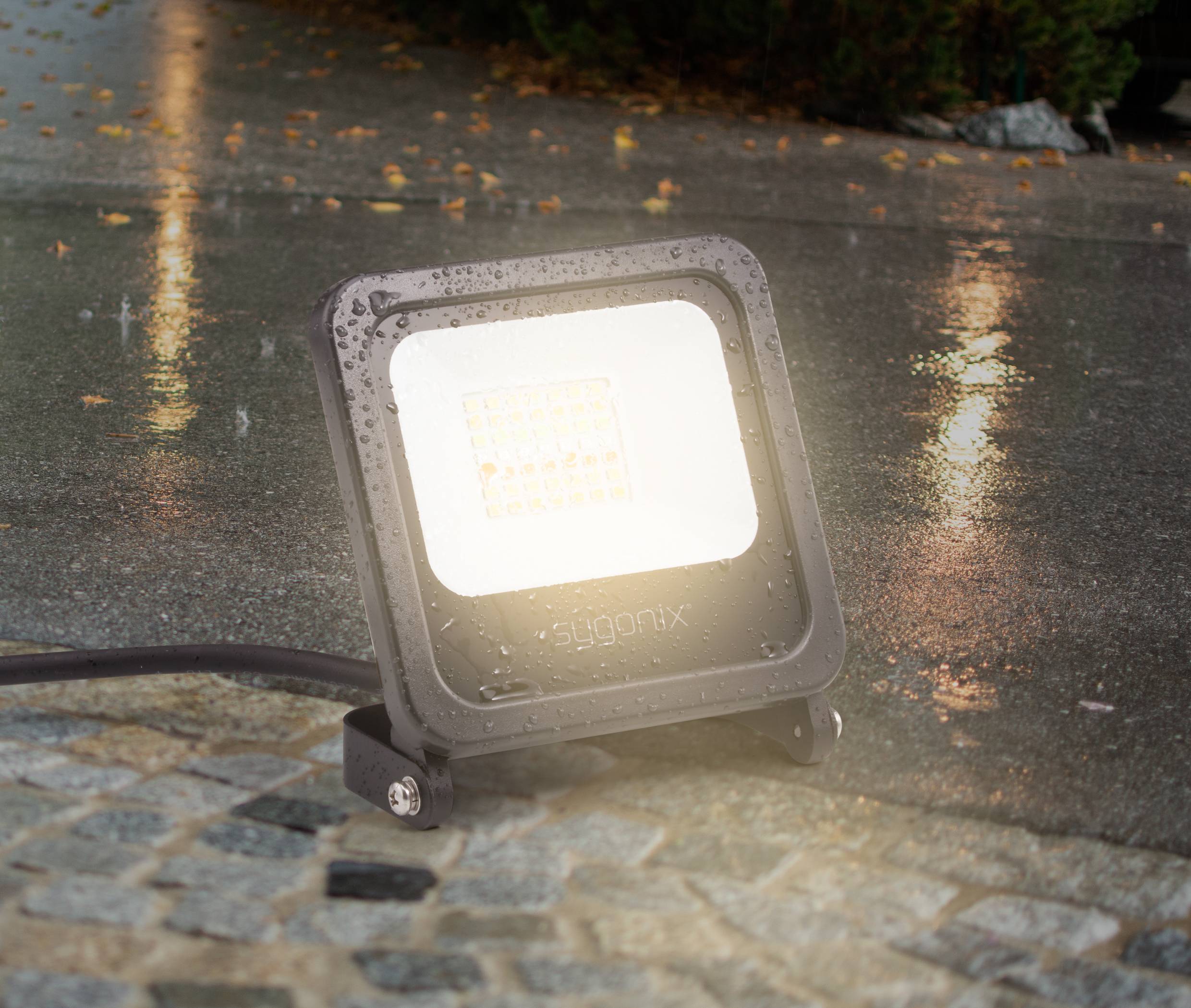 A waterproof LED floodlight illuminates the wet cobblestones. Rain reflects on the ground, lighting emphasising the moisture.