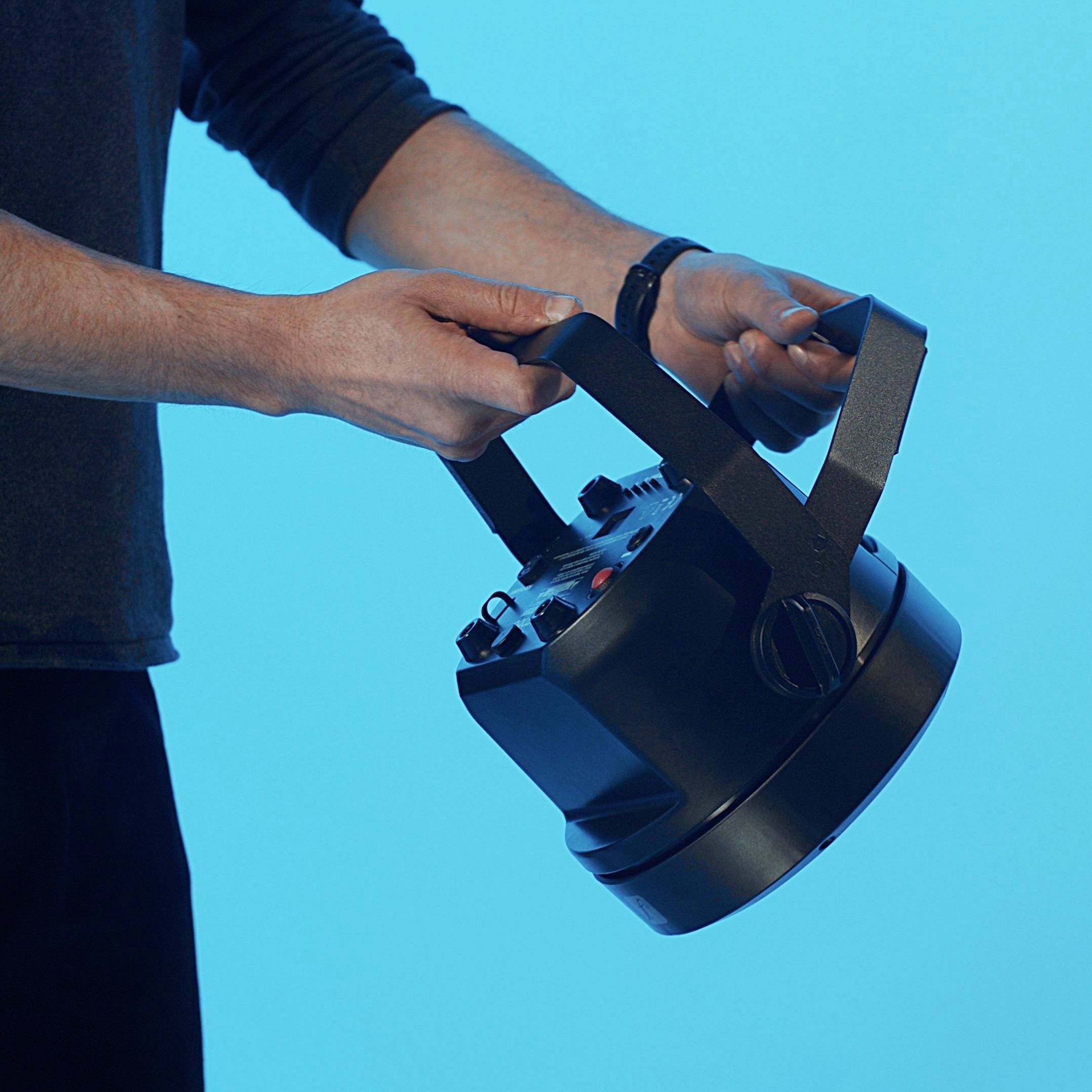 A person holds a stage spotlight against a blue background. The person is wearing a dark long-sleeved shirt.