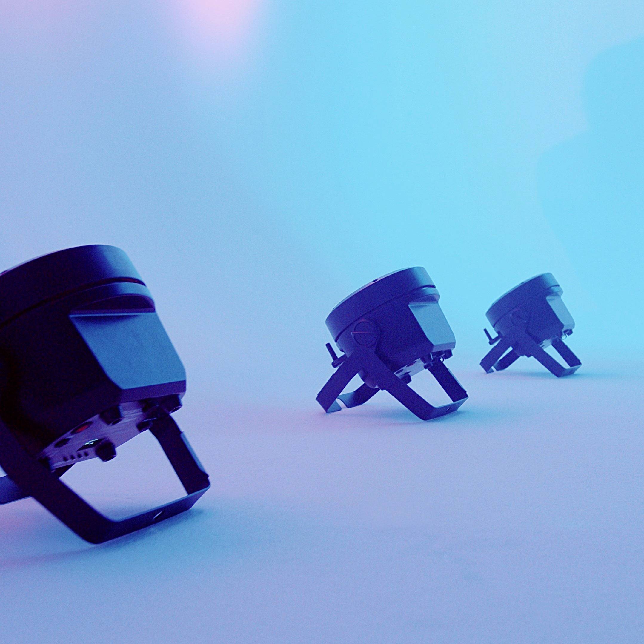 Three black spotlights are projecting blue light on a stage. They are diagonally positioned in the space, rising to the right.