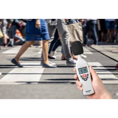 A person is holding a sound level meter displaying 91.9 decibels, with people crossing a bustling street in the background.