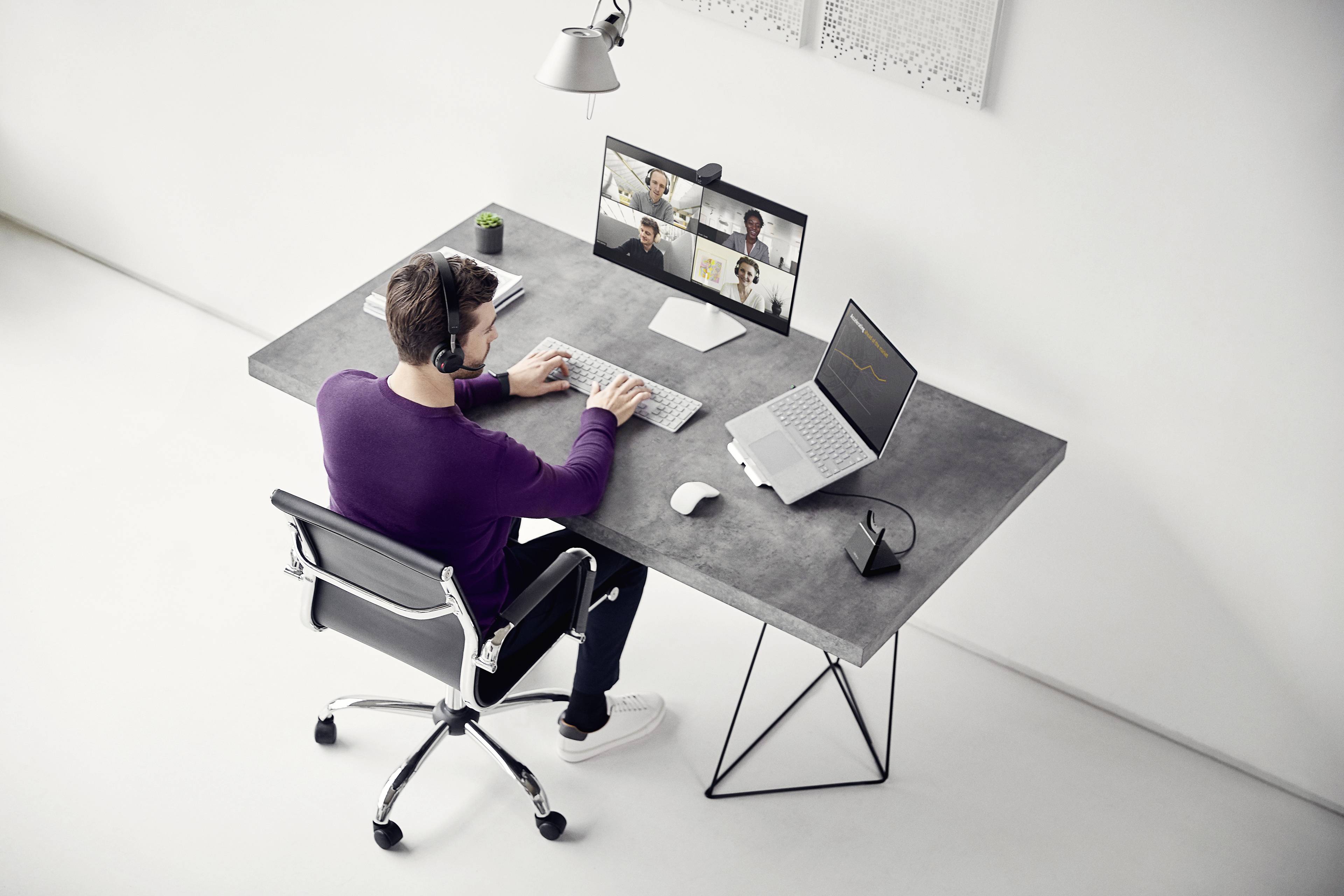 A man is sitting at a modern desk and participating in a video conference on a laptop. He is wearing headphones.