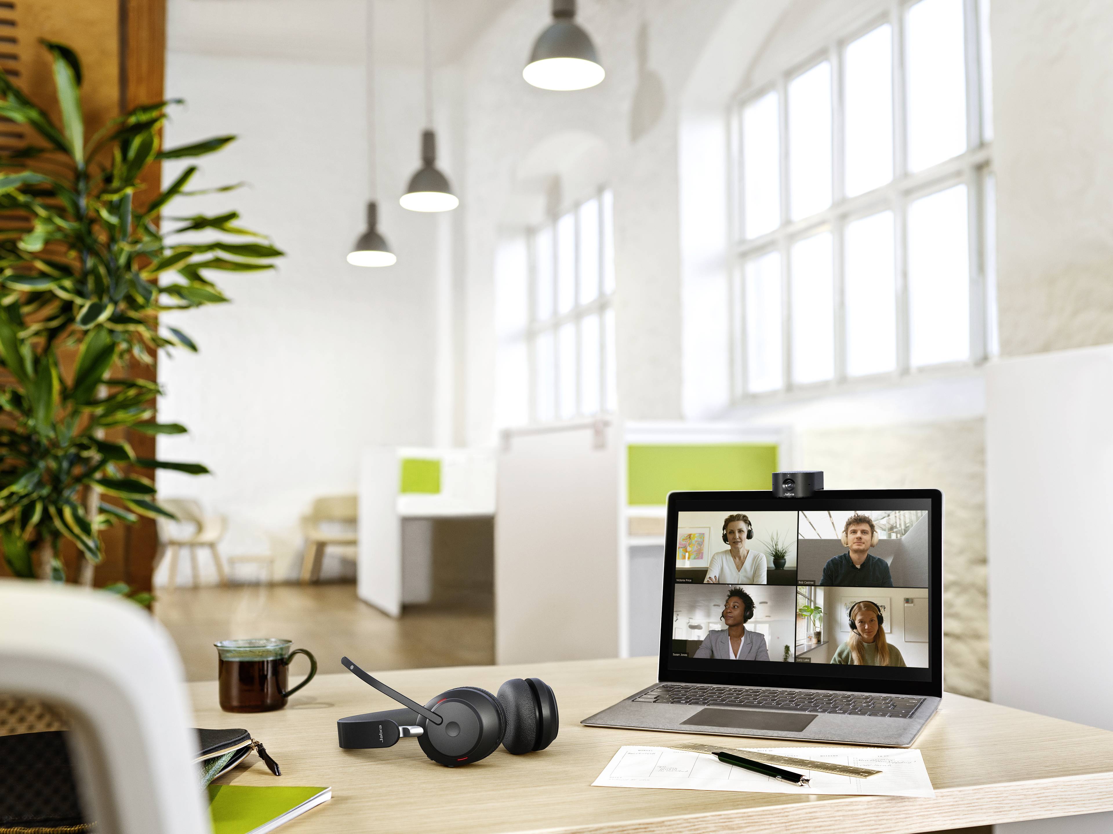 Laptop on desk with video call open. Four people on the screen. Office in the background. Headset, notes and drink beside it.
