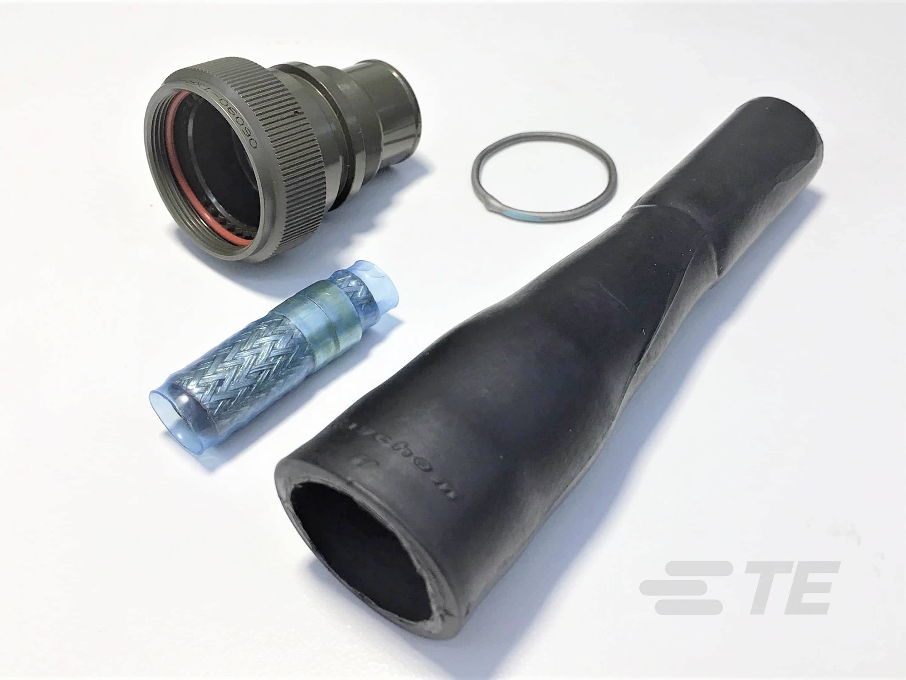 A military connector, a sealing rubber, a bluish casing and a cable protection are lying on a white surface.
