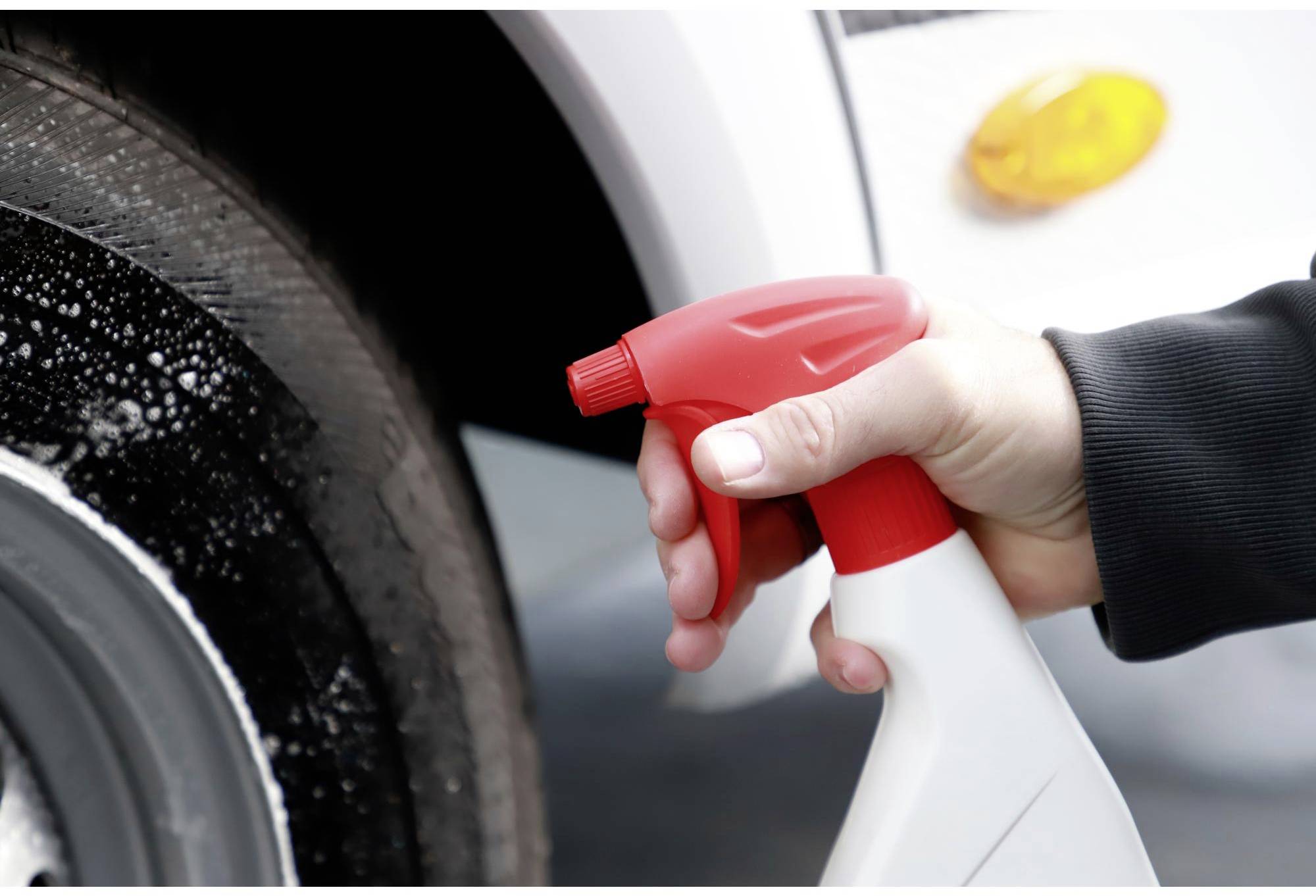 A hand is holding a red spray bottle and spraying cleaning solution onto a car tyre. A white vehicle can be seen in the background.