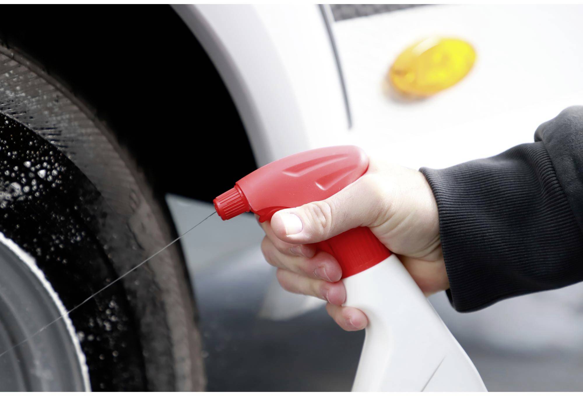 A hand holds a spray bottle and sprays cleaning solution onto a vehicle's wheel to generate foam on the tyre.