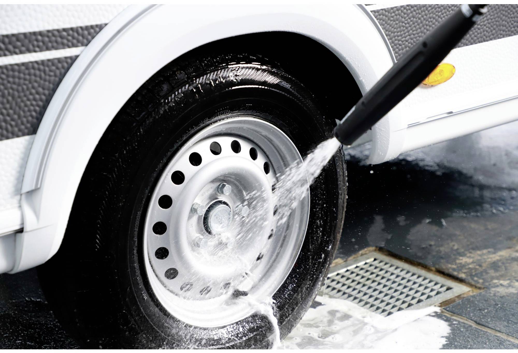 A pressure washer sprays water onto a car tyre to remove dirt. The tyre has a silver wheel with several holes.