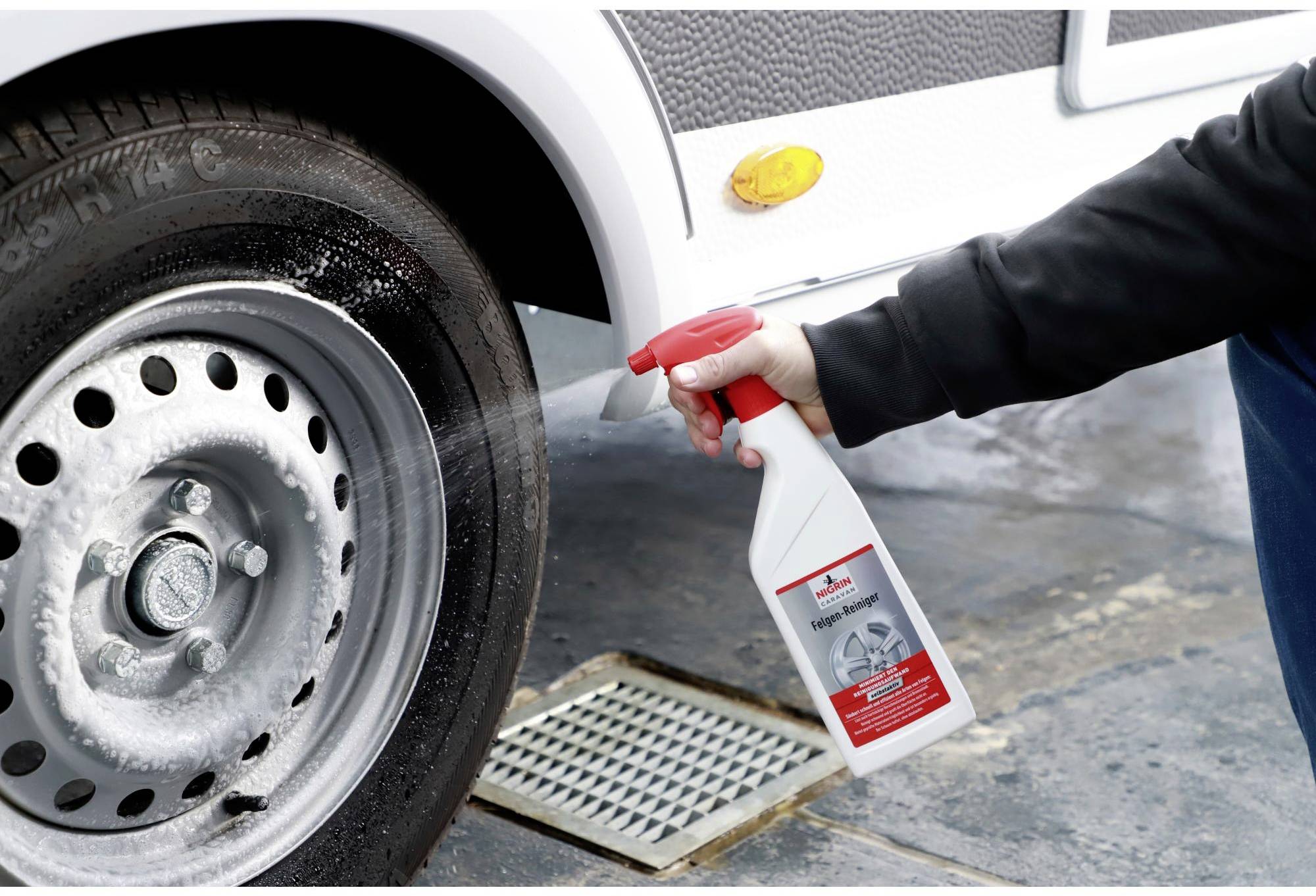 A hand sprays cleaning solution onto a car tyre wheel. The tyre is mounted on a vehicle. The ground is wet from spraying.