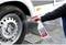 A hand sprays cleaning solution onto a car tyre wheel. The tyre is mounted on a vehicle. The ground is wet from spraying.