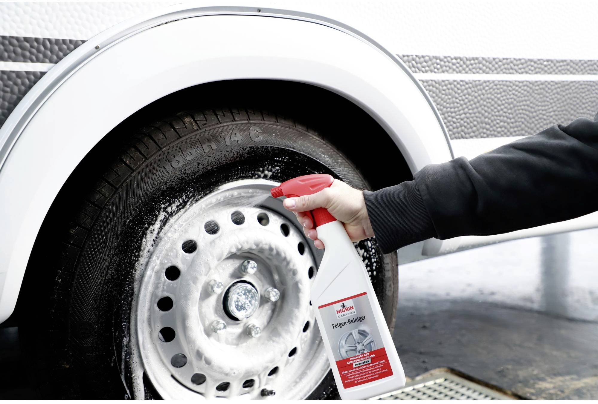 A hand sprays a cleaning product onto a dirty car tyre. The cleaner generates white foam on the wheel rim.