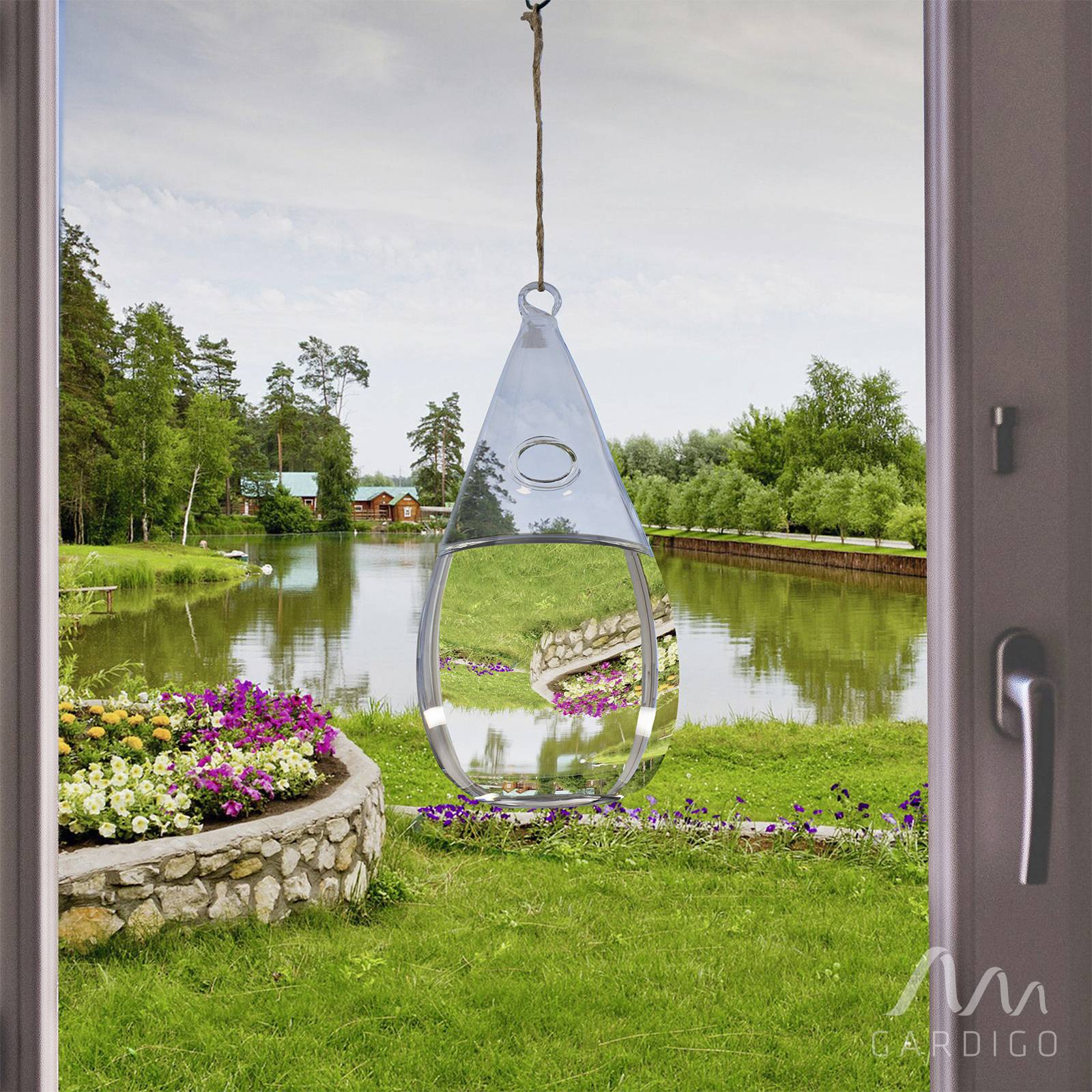 View from a window: A hanging water droplet mirror reflects a landscape with a lake, flowers, trees and a wooden house in the background.