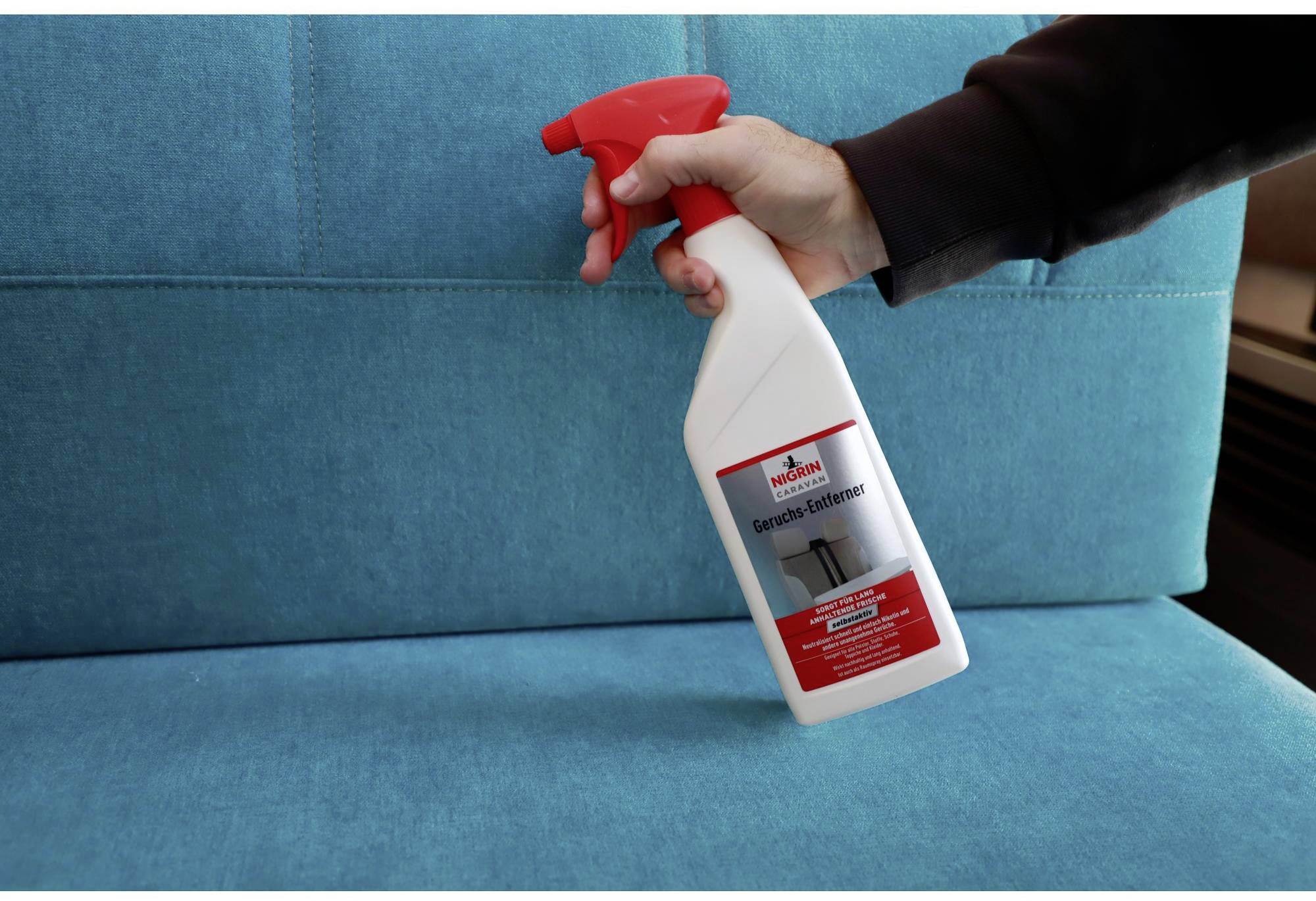 A hand holds an odour neutraliser spray over a blue sofa cushion.