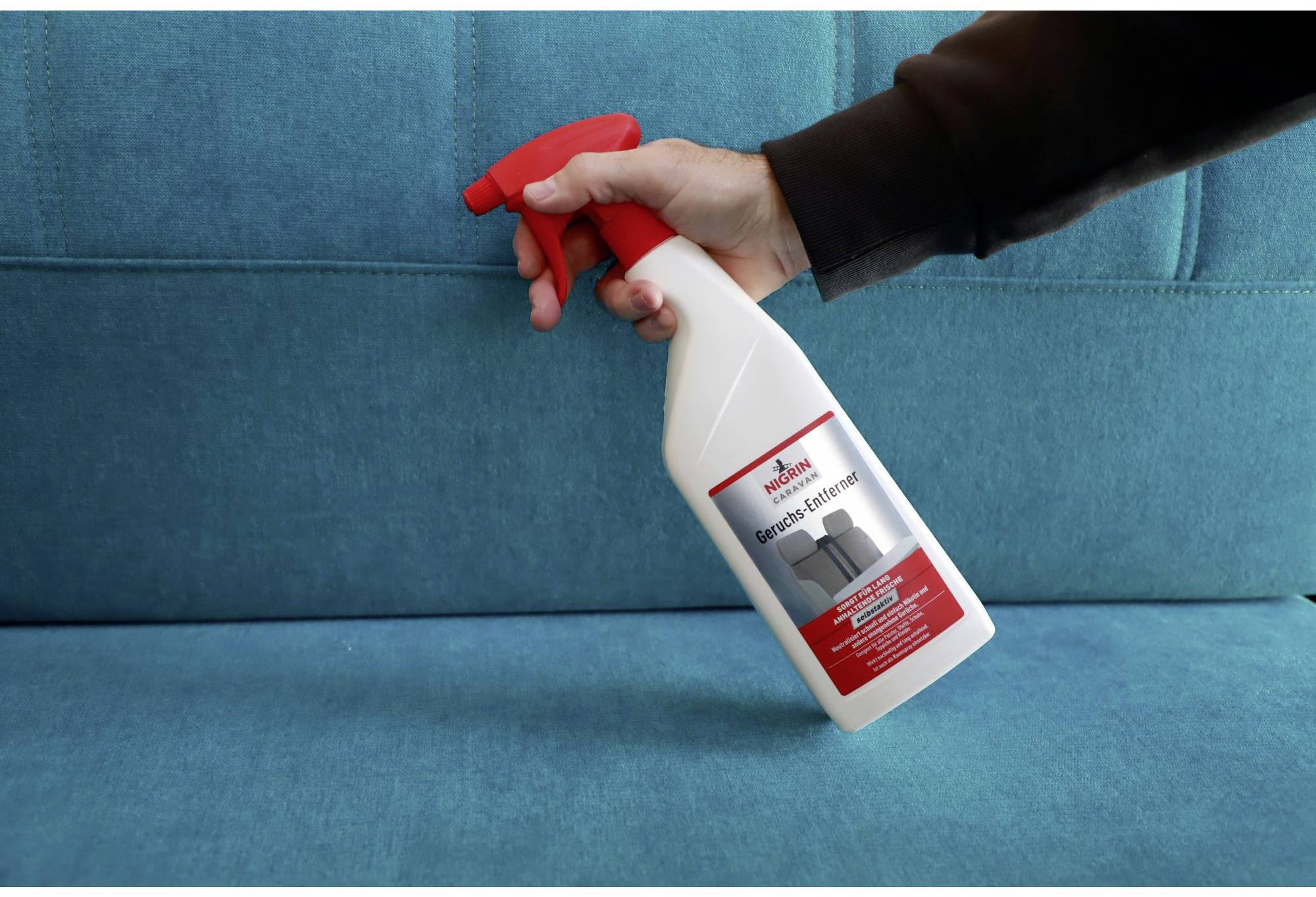 A hand is holding a bottle of upholstery cleaner over a blue sofa.