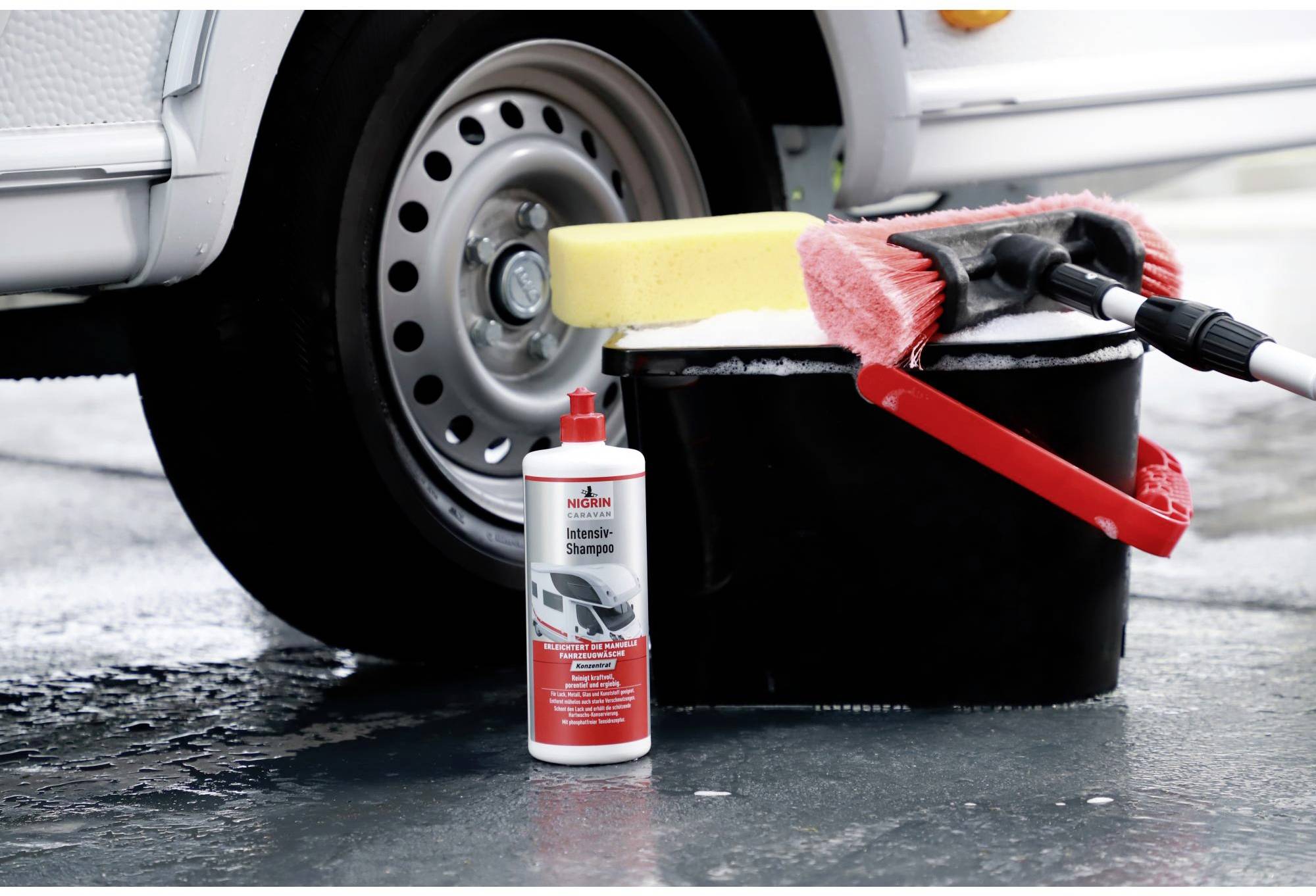 A car stands on wet asphalt. Beside it are buckets with a sponge and brush, along with a bottle of car shampoo. The scene shows car cleaning accessories.