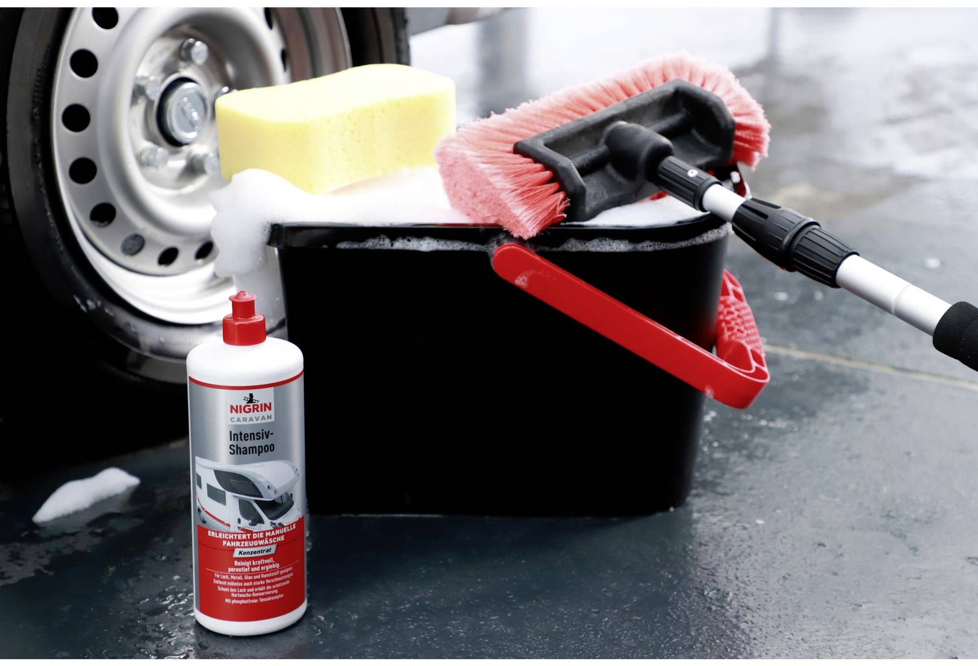 'Car cleaning set with shampoo bottle, black bucket, red wash mop, yellow sponge; tyres in the background.'