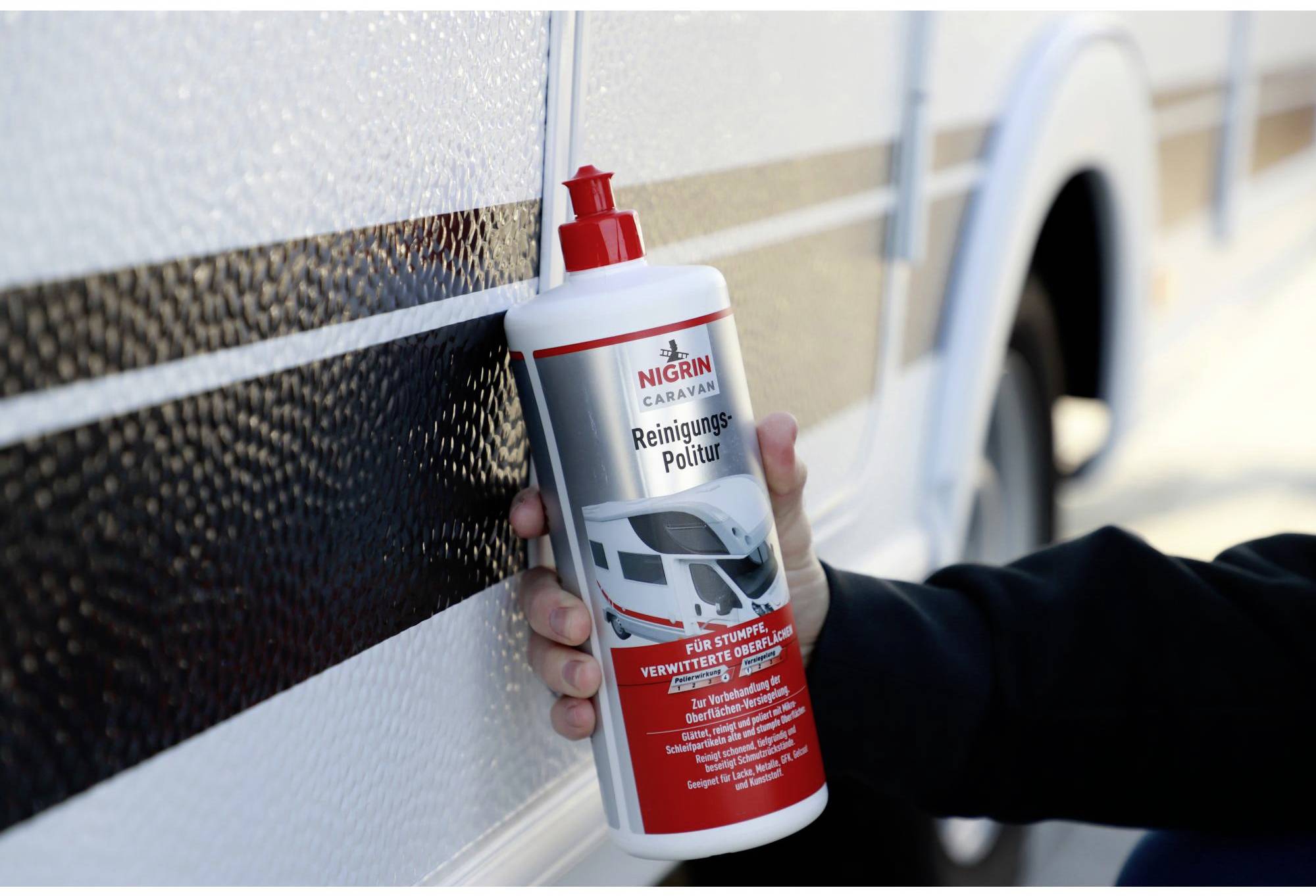 A person is holding a bottle of NIGRIN Caravan Cleaner against the exterior wall of a motorhome.