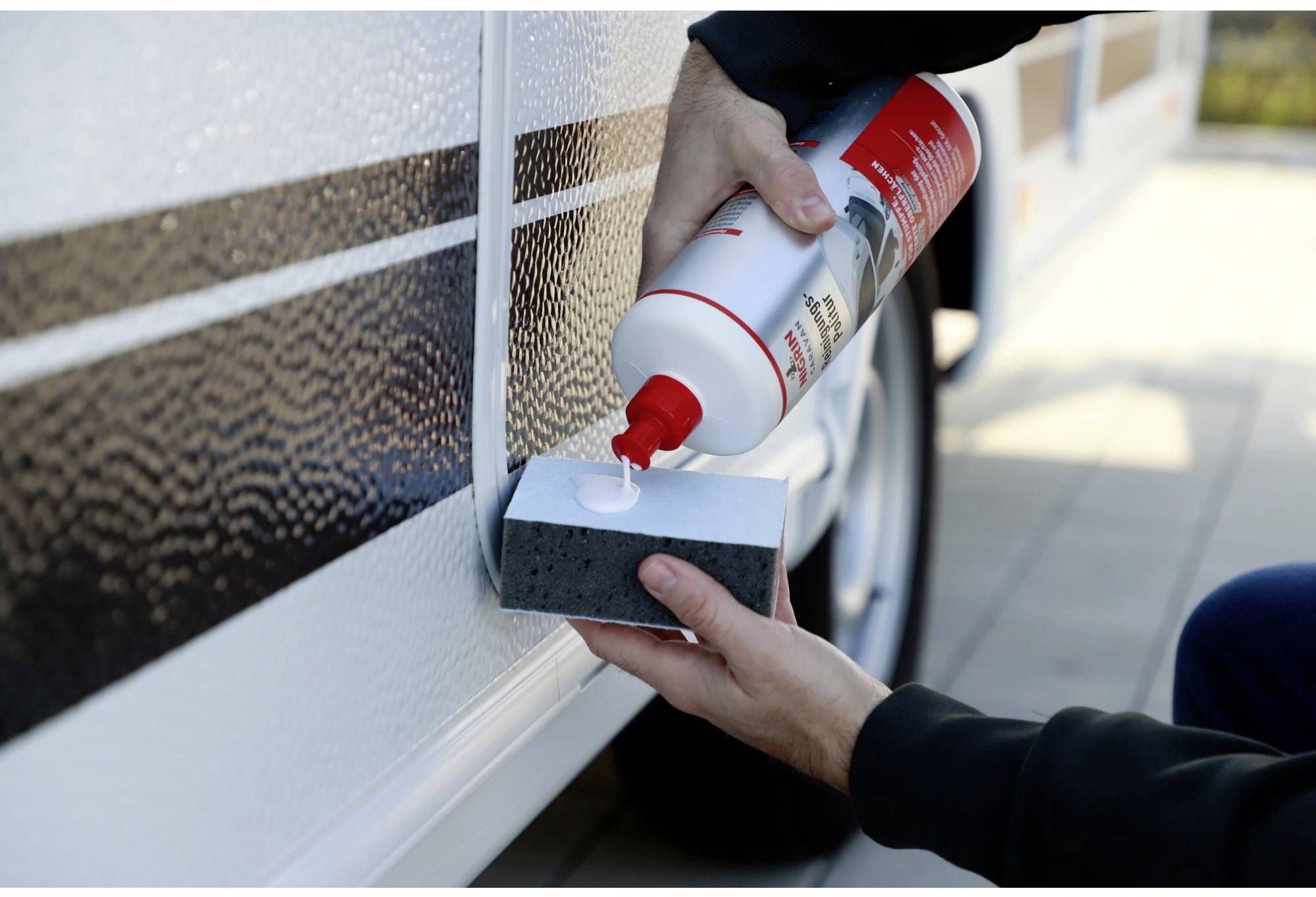 A person is applying cleaning solution to the exterior surface of a vehicle using a sponge. The vehicle is outdoors.
