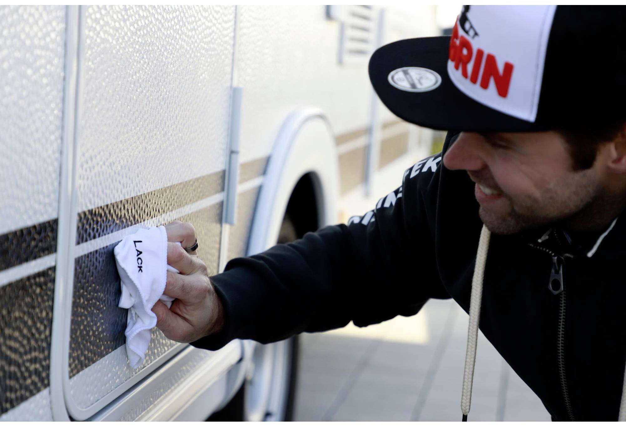 A man is cleaning the exterior surface of a campervan with a cloth. He is wearing a black cap and smiling while working.