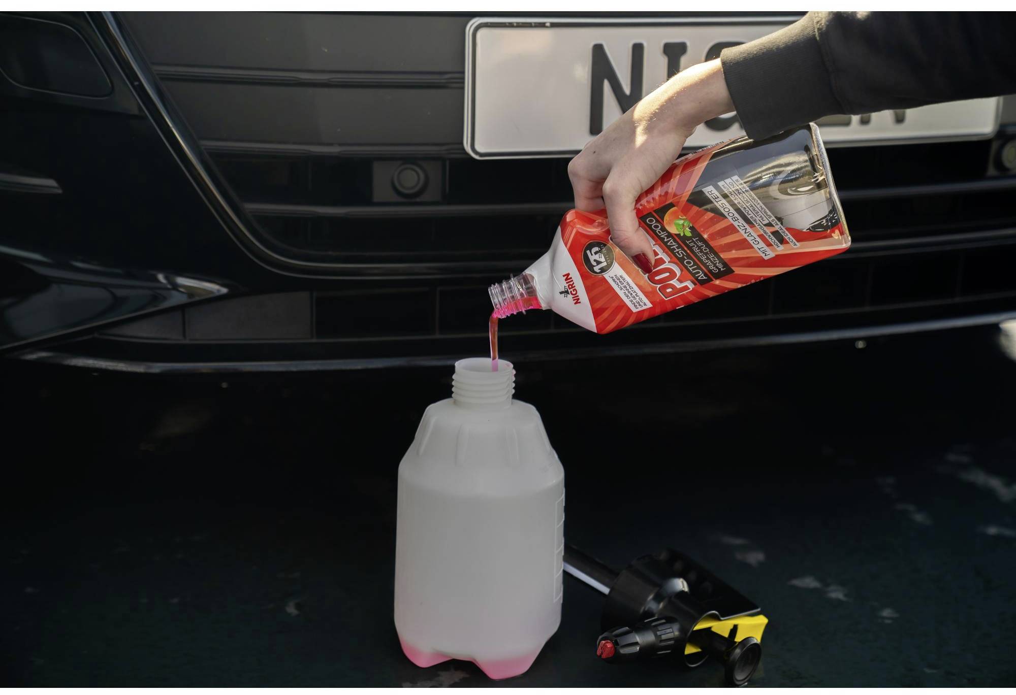 A person is pouring pink liquid from a container into a large container under the bonnet of a black car.