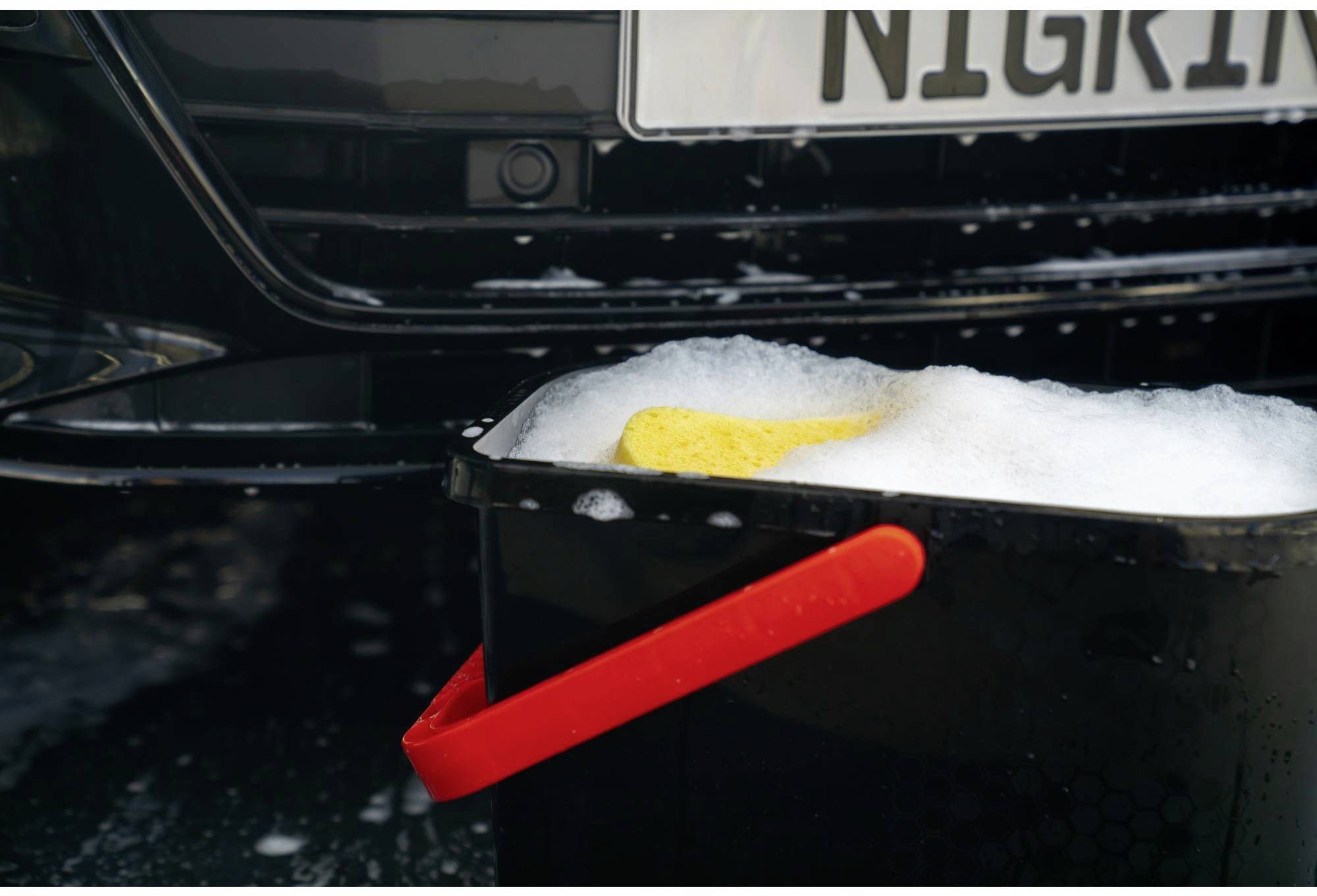A black bucket with soapy water and a yellow sponge is standing in front of a black car with a partially visible number plate.