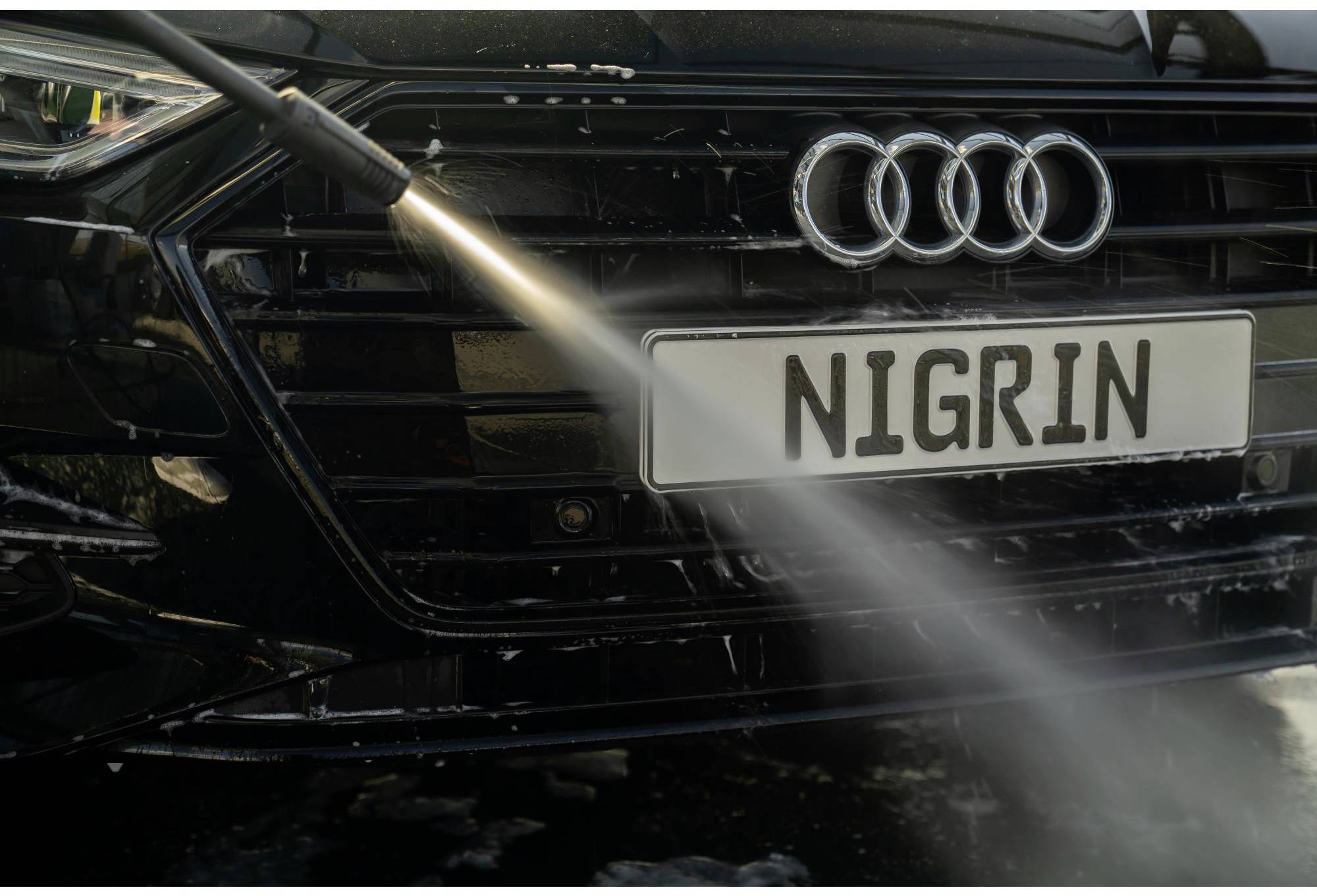 A black car is being cleaned with a pressure washer. The focus is on the radiator grille and the headlight.