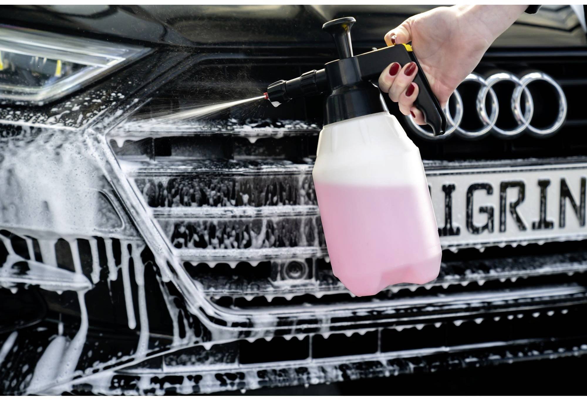 A hand sprays a pink liquid onto the front of a car using a cleaning device, which is covered in foam.